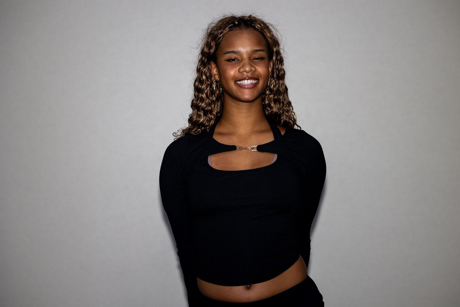 A cheerful young woman with curly blonde-highlighted hair smiles warmly in a black cutout crop top.