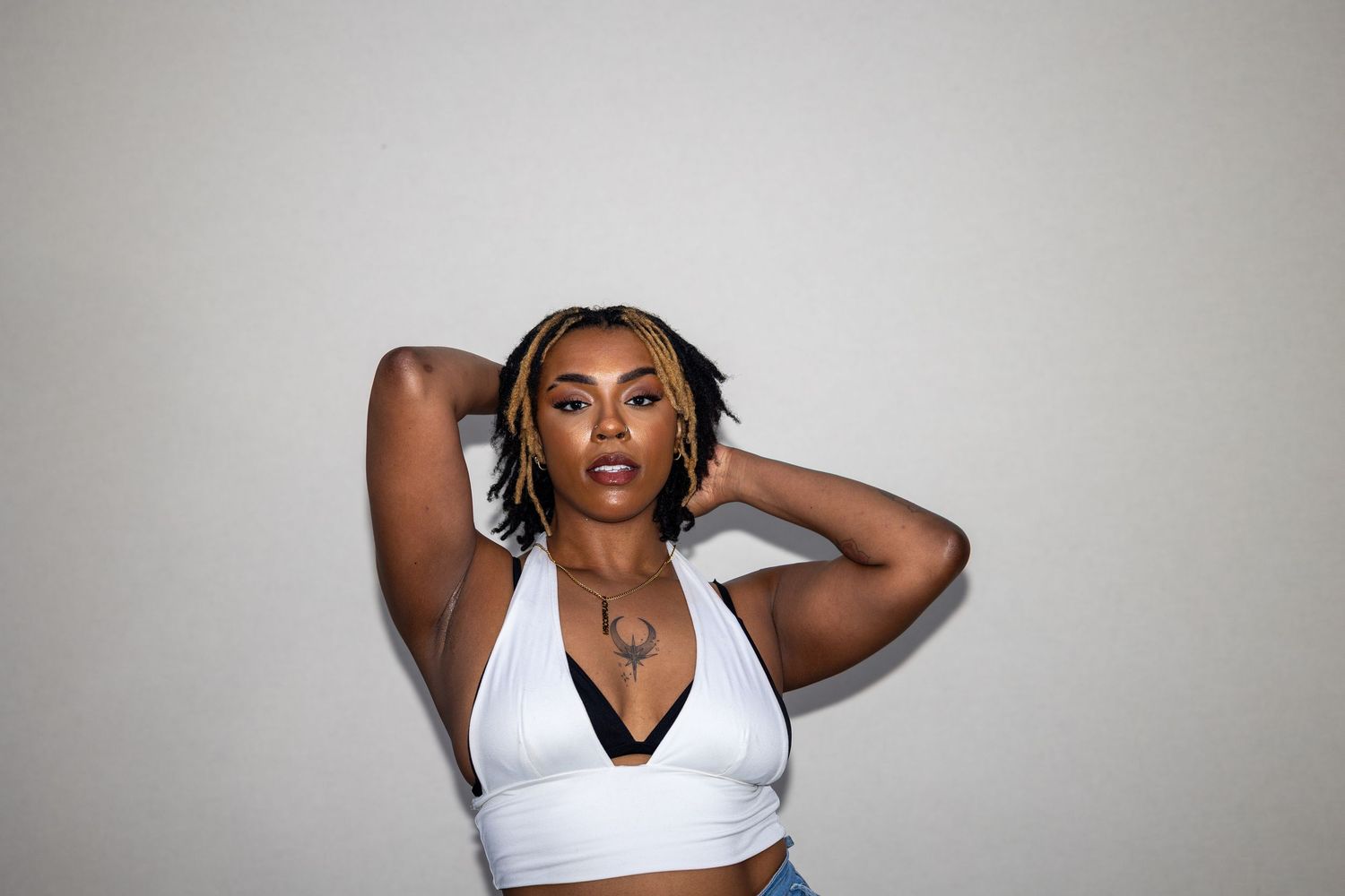 Woman in white halter crop top with hands behind head poses gracefully against light background.