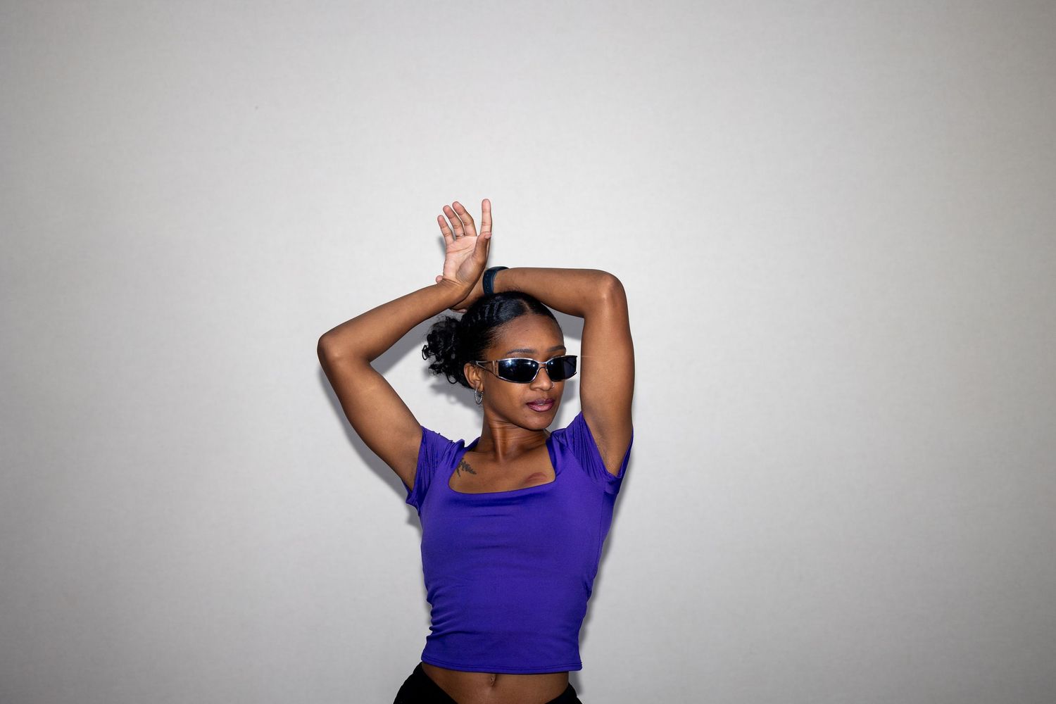 Stylish Black woman in purple crop top and sunglasses dancing with arms raised overhead.