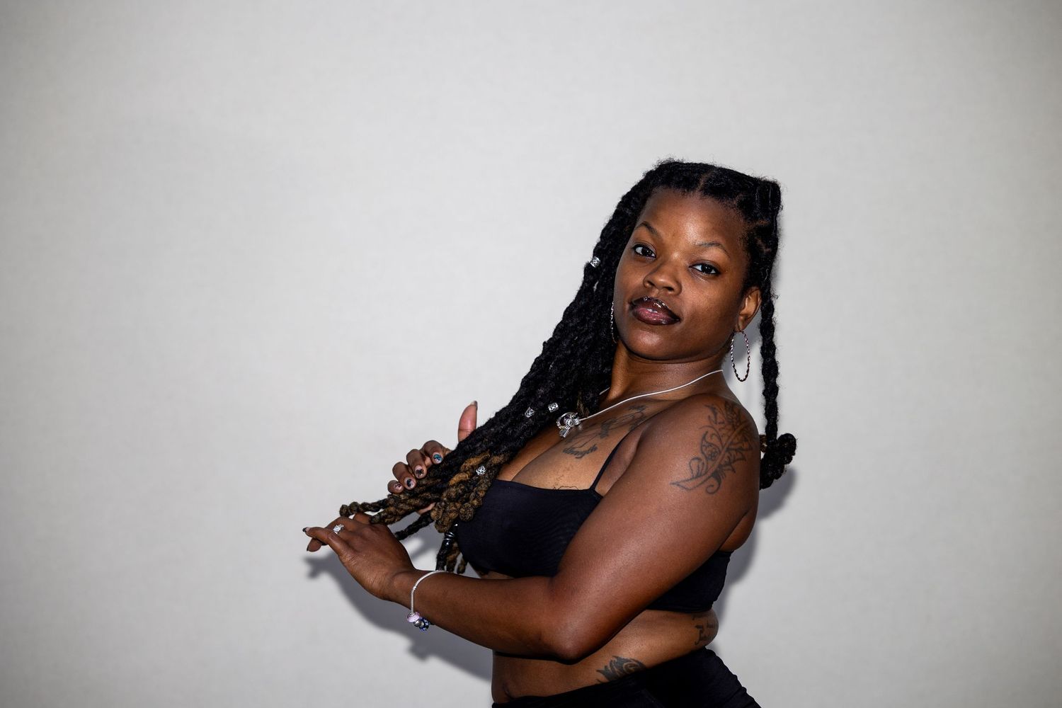Young woman with braided locs poses holding a flute, wearing a black crop top with a tattoo visible on her arm.