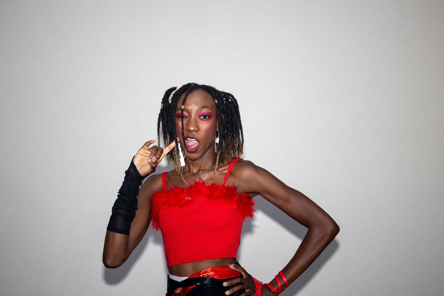 Woman in red feathered crop top and braids poses playfully against white background.