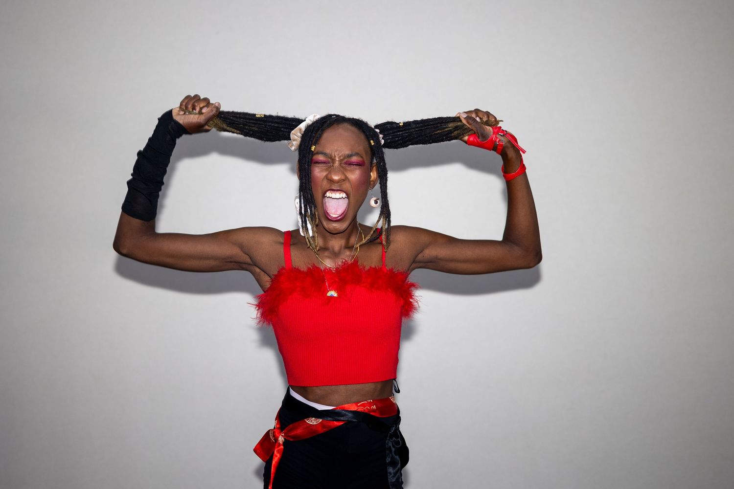 A woman in a red feathered crop top pulls her braids up while sticking out her tongue playfully.