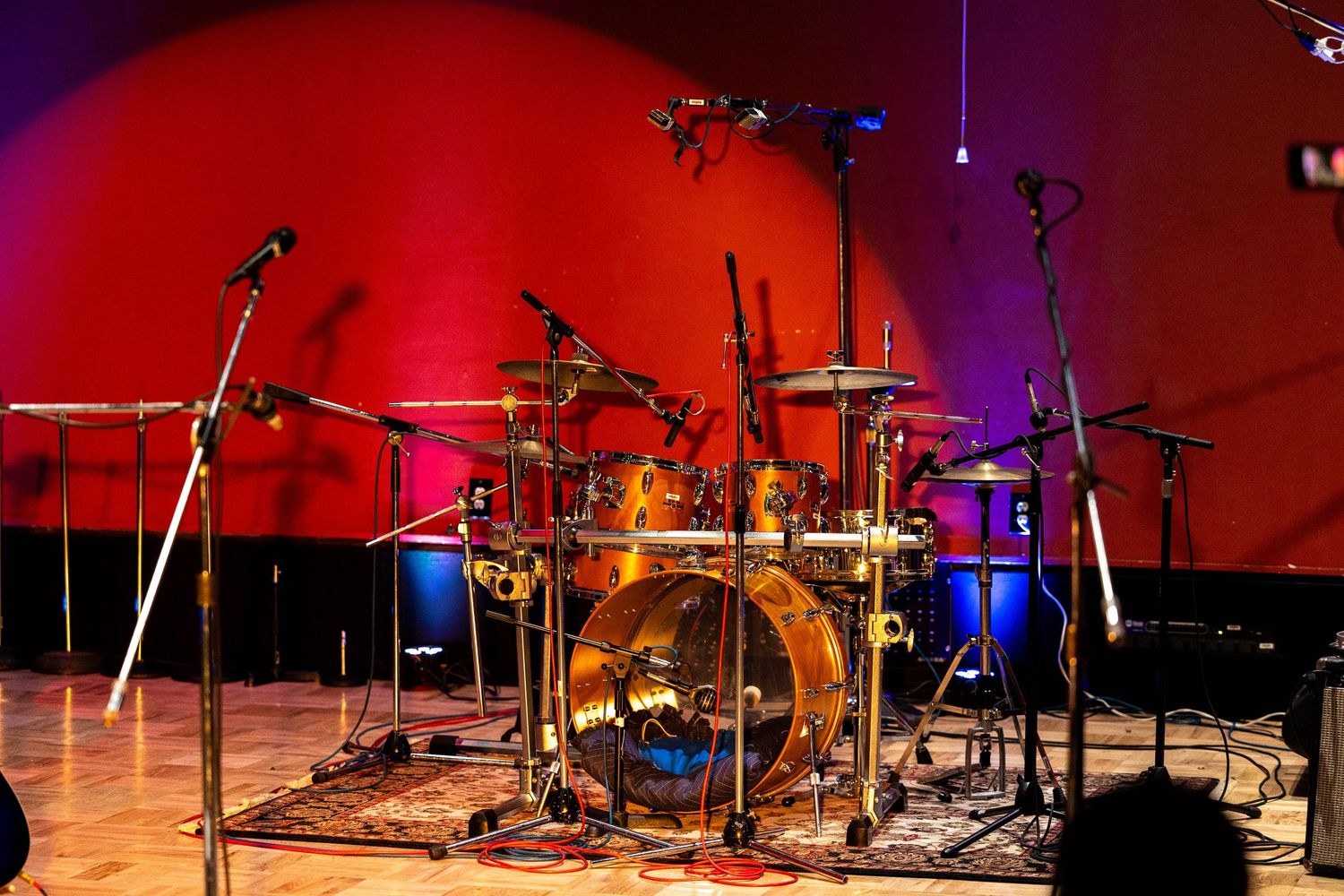 A professional drum kit set up on a concert stage with microphones, red and blue lighting backdrop.