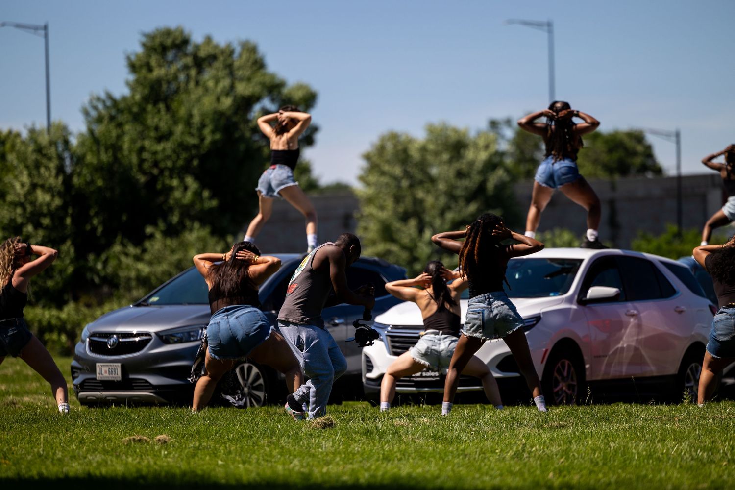 Group of women performing a choreographed dance routine around parked cars on a sunny day.