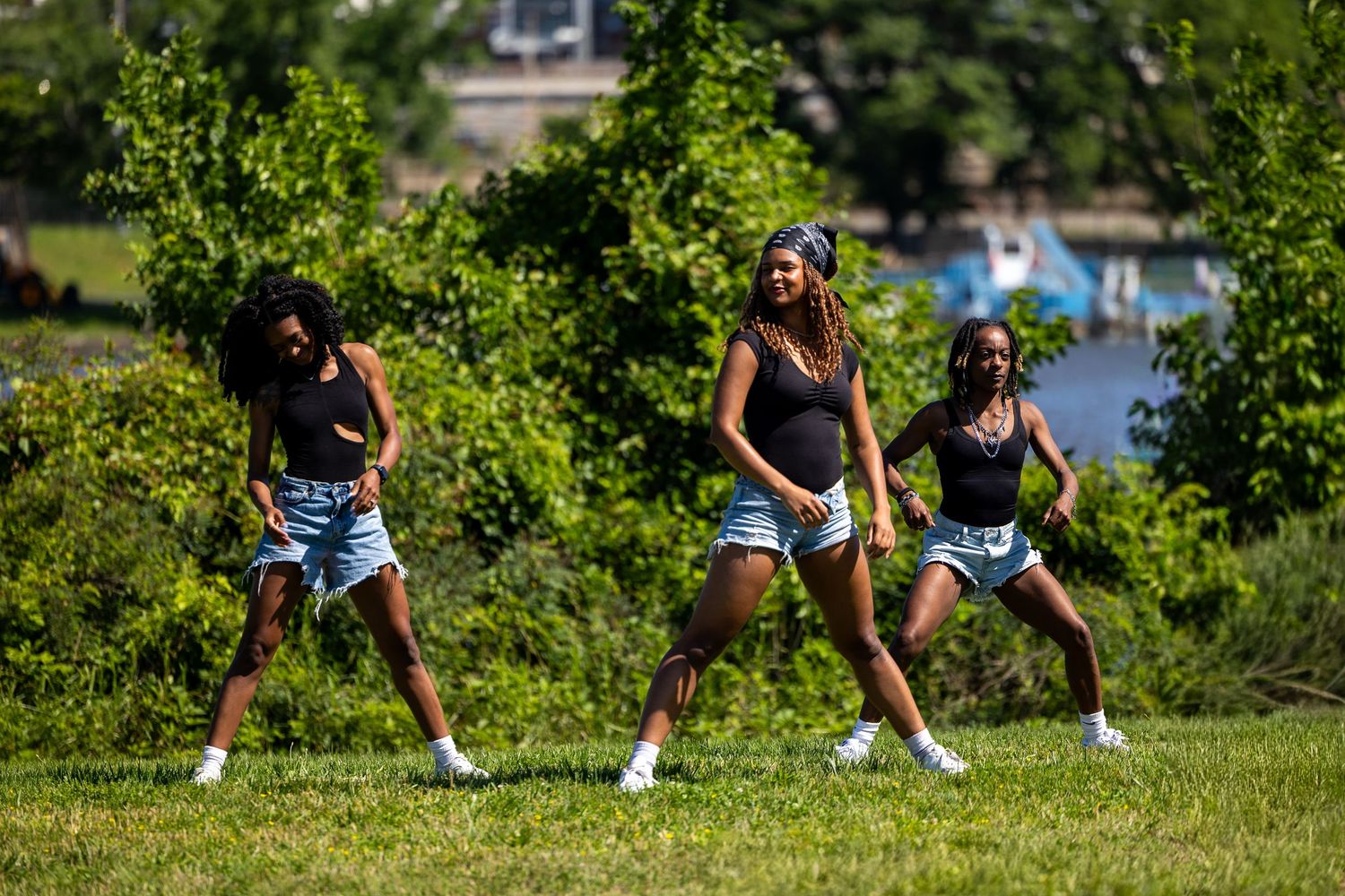 Three young women dancing in sync outdoors on grass near a waterfront with lush green trees.