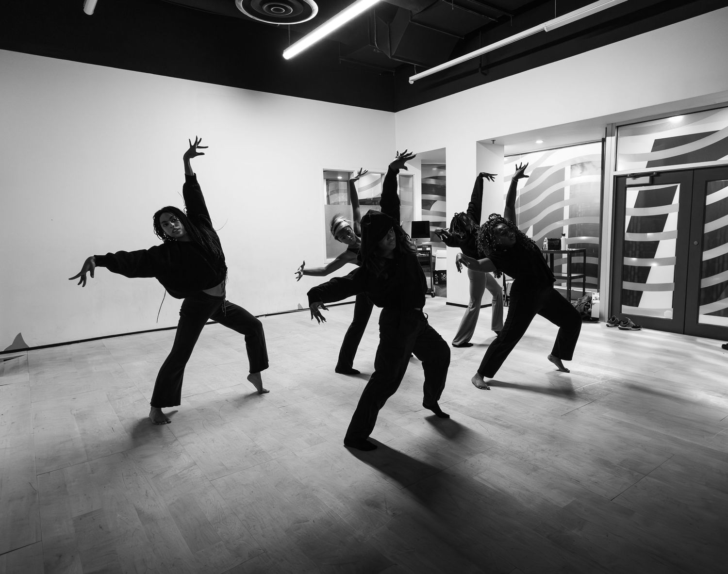 Four dancers in black perform dynamic poses in a modern dance studio, captured in black and white photography.