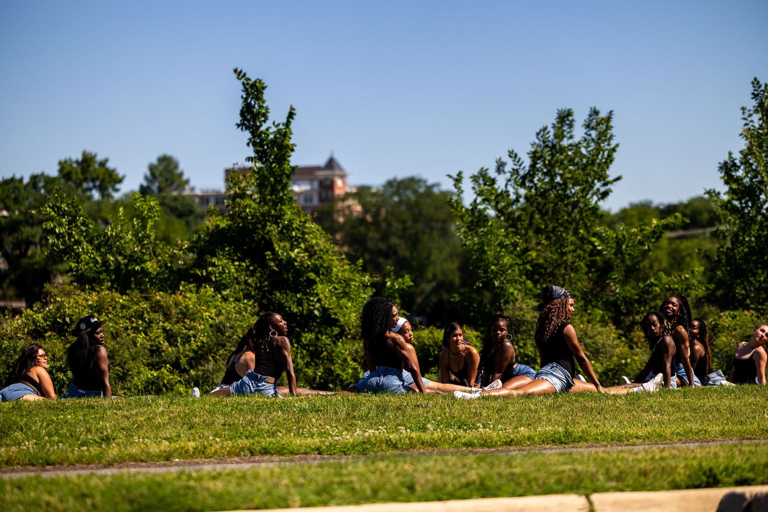 Group of young women relaxing on green grass in a sunny park with trees and a building in the background.