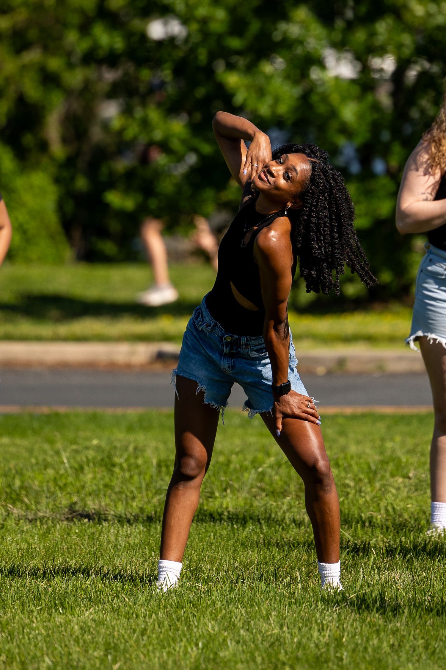 Young Black woman dancing outdoors in denim shorts and black crop top on green grass in bright sunlight.
