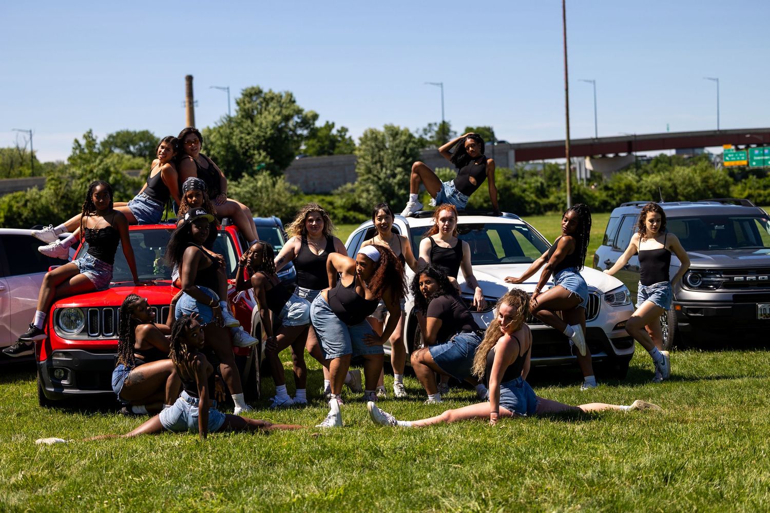 Group of young women posing together outdoors near cars on a sunny day.
