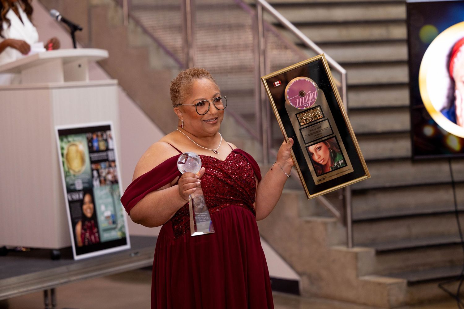 Woman in red gown holds award plaque at formal ceremony event.