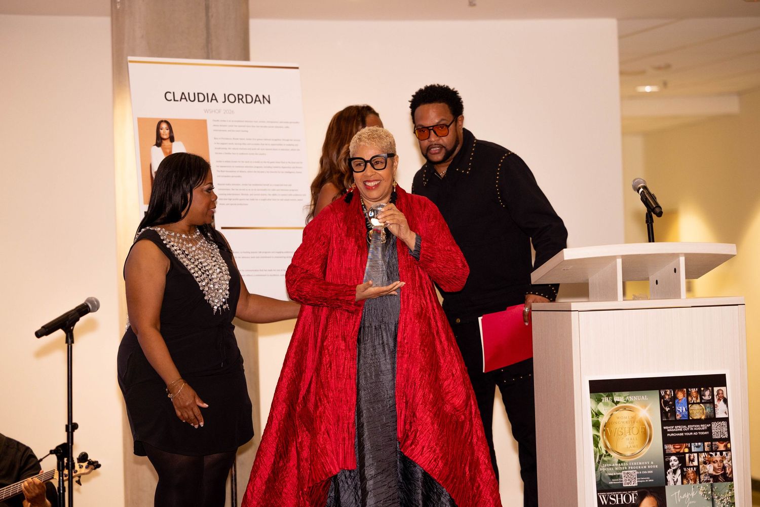 Elegantly dressed woman in red coat speaks at podium flanked by two guests at gala.