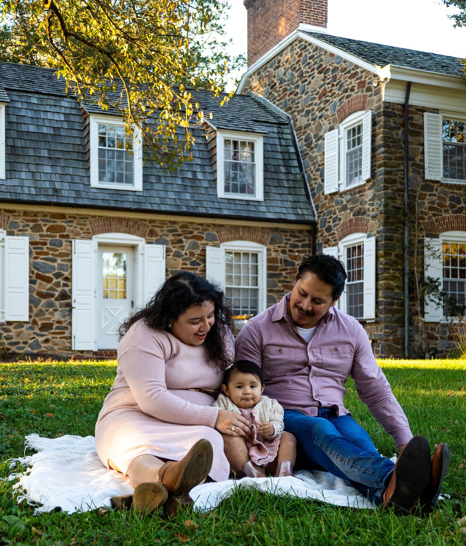Family of three sitting on blanket in front of stone colonial house on green lawn.