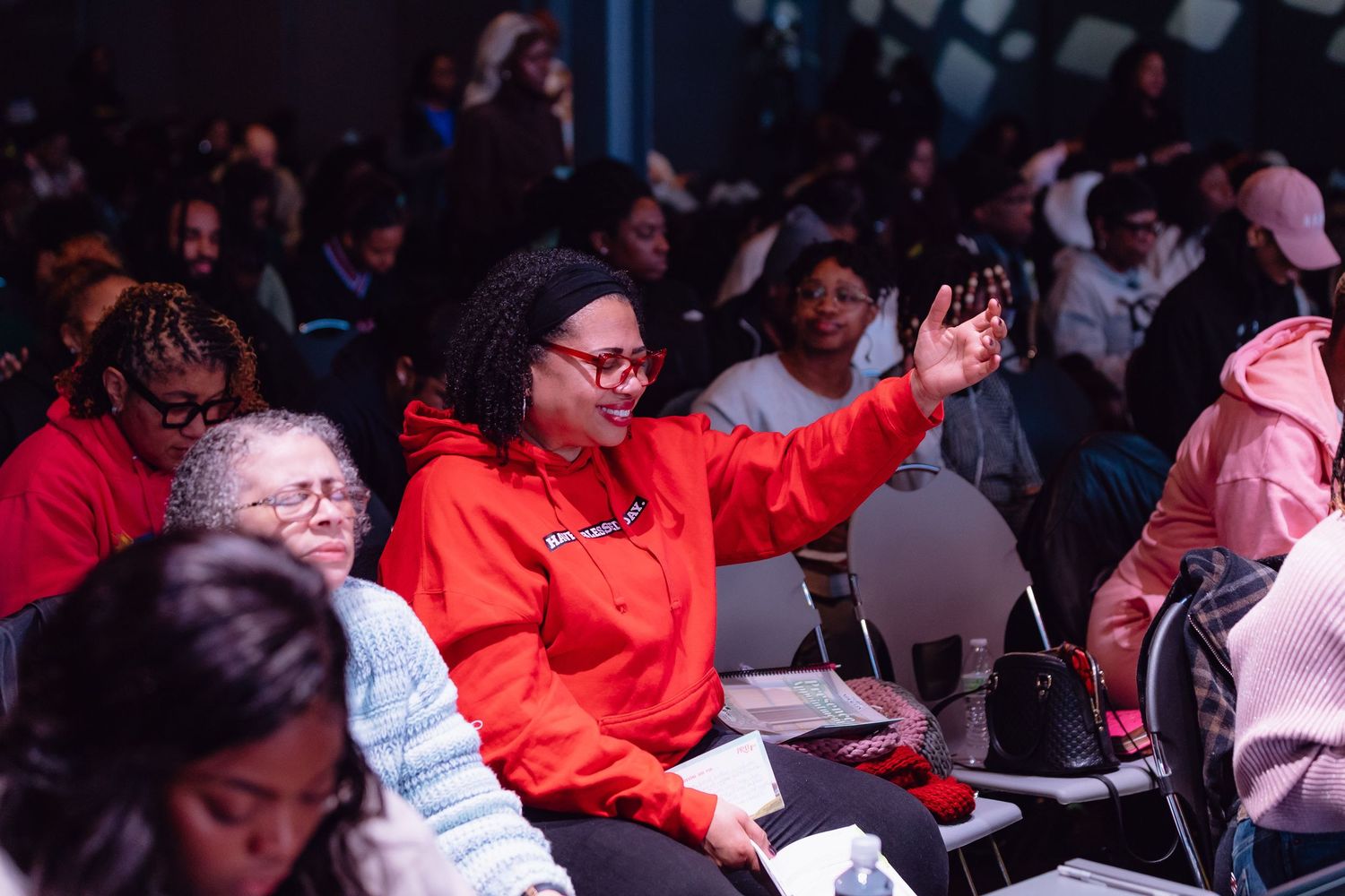 A crowd of people in red and casual attire sit in an auditorium with someone raising their hand during an event.