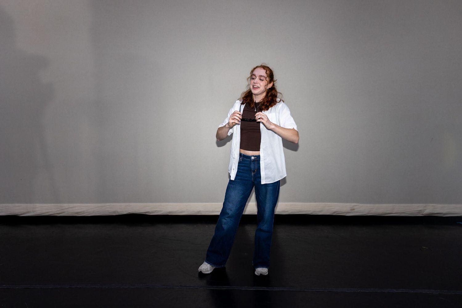 A young woman in jeans and white shirt gestures expressively while performing on a dark studio stage.