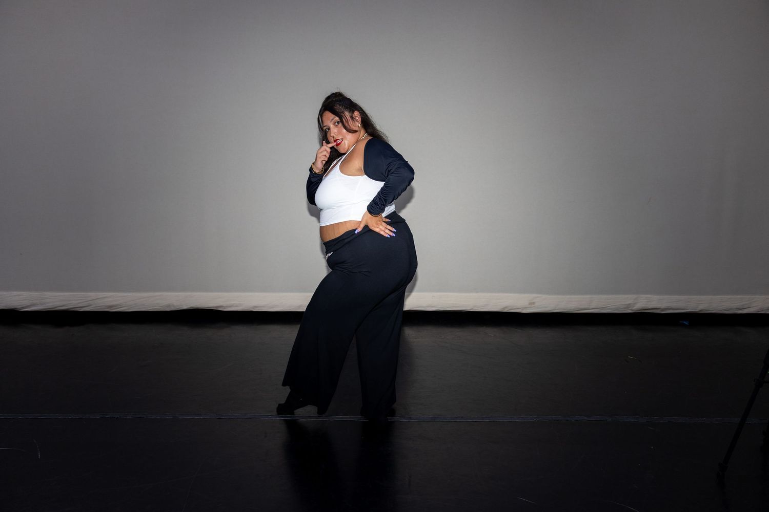A dancer poses dramatically in a white crop top and navy wide-leg pants against a gray studio backdrop.