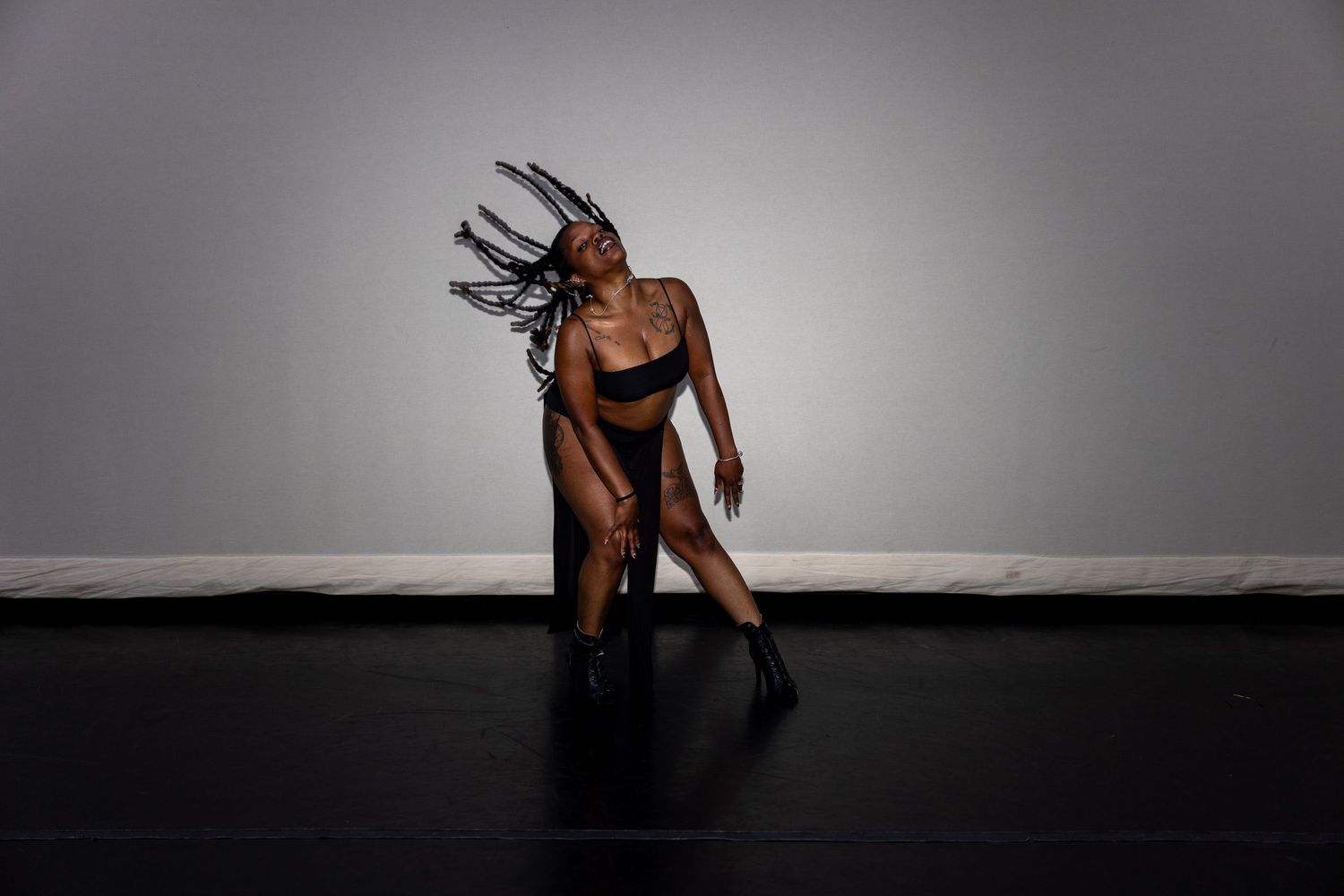A dancer in black tosses braids back in a dynamic pose on a dark studio floor.