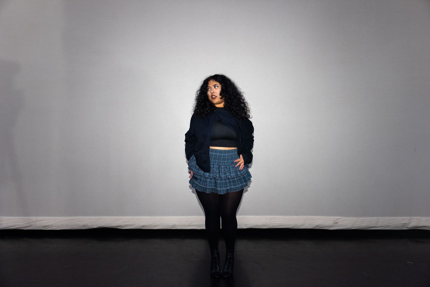 A woman with curly hair poses in a black crop top, plaid mini skirt, and tights against a gray background.