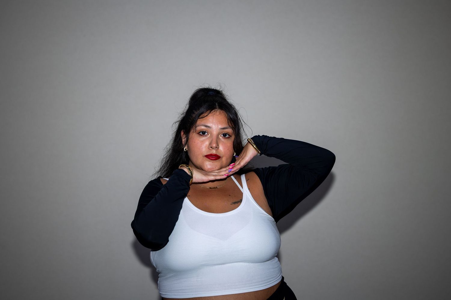 A plus-size woman in a white sports bra poses confidently with hands near her face against a gray backdrop.