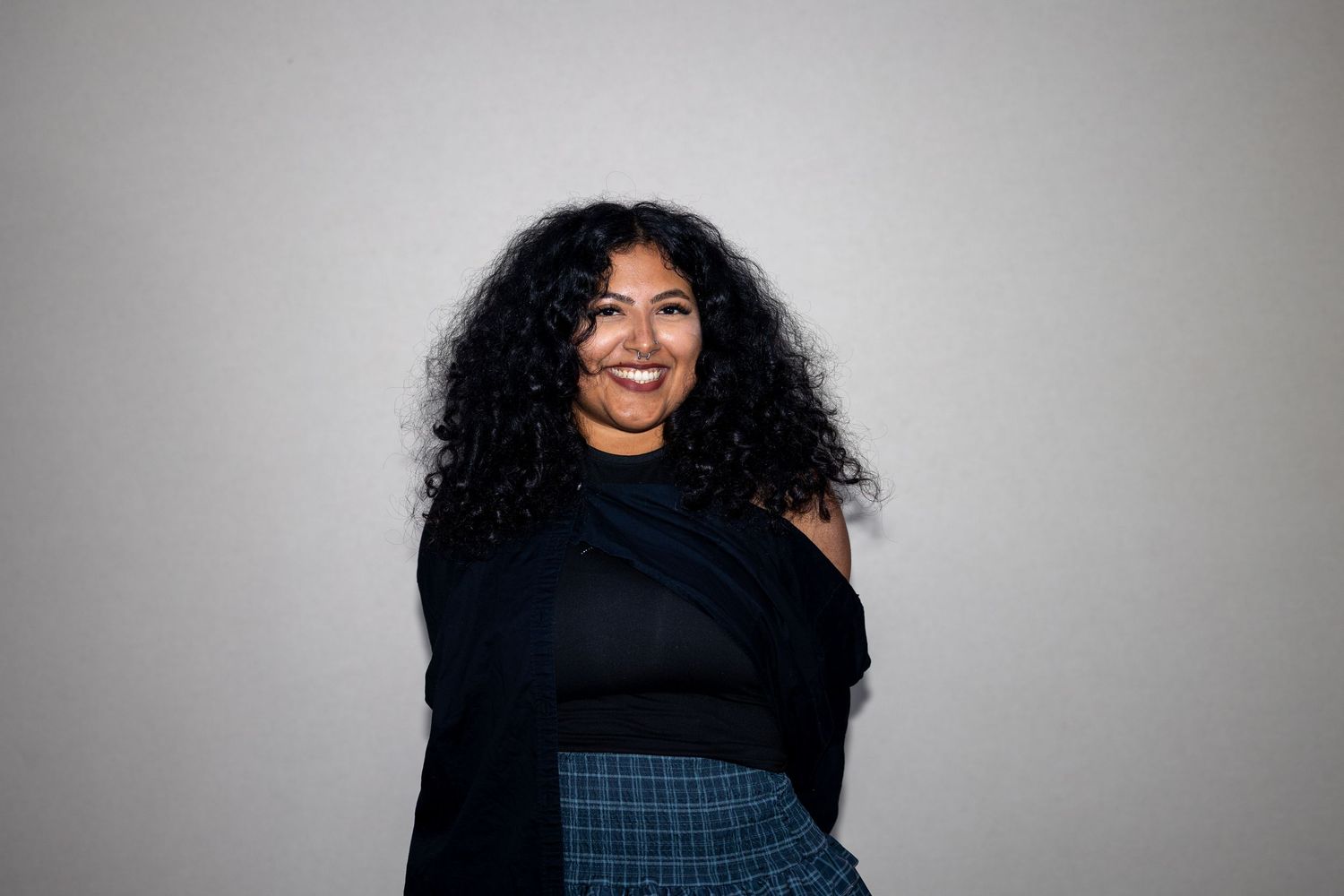 Woman with voluminous curly hair wearing black off-shoulder top and plaid skirt smiles warmly.