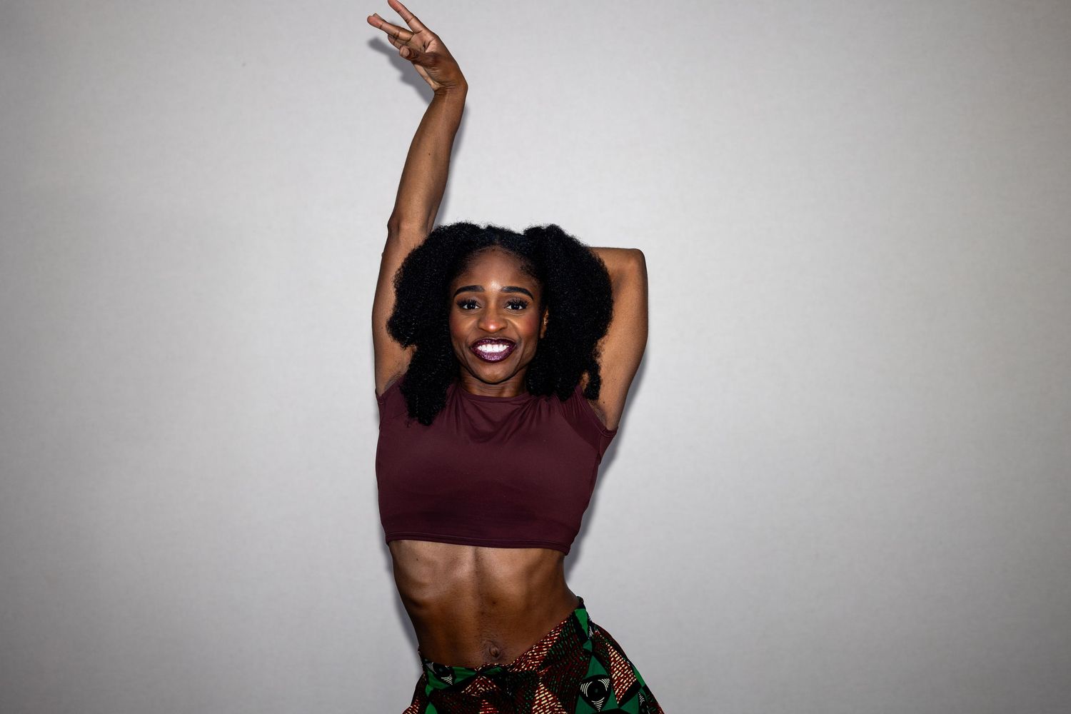 A fit woman in a burgundy crop top and floral skirt jumps joyfully with a peace sign against a gray background.