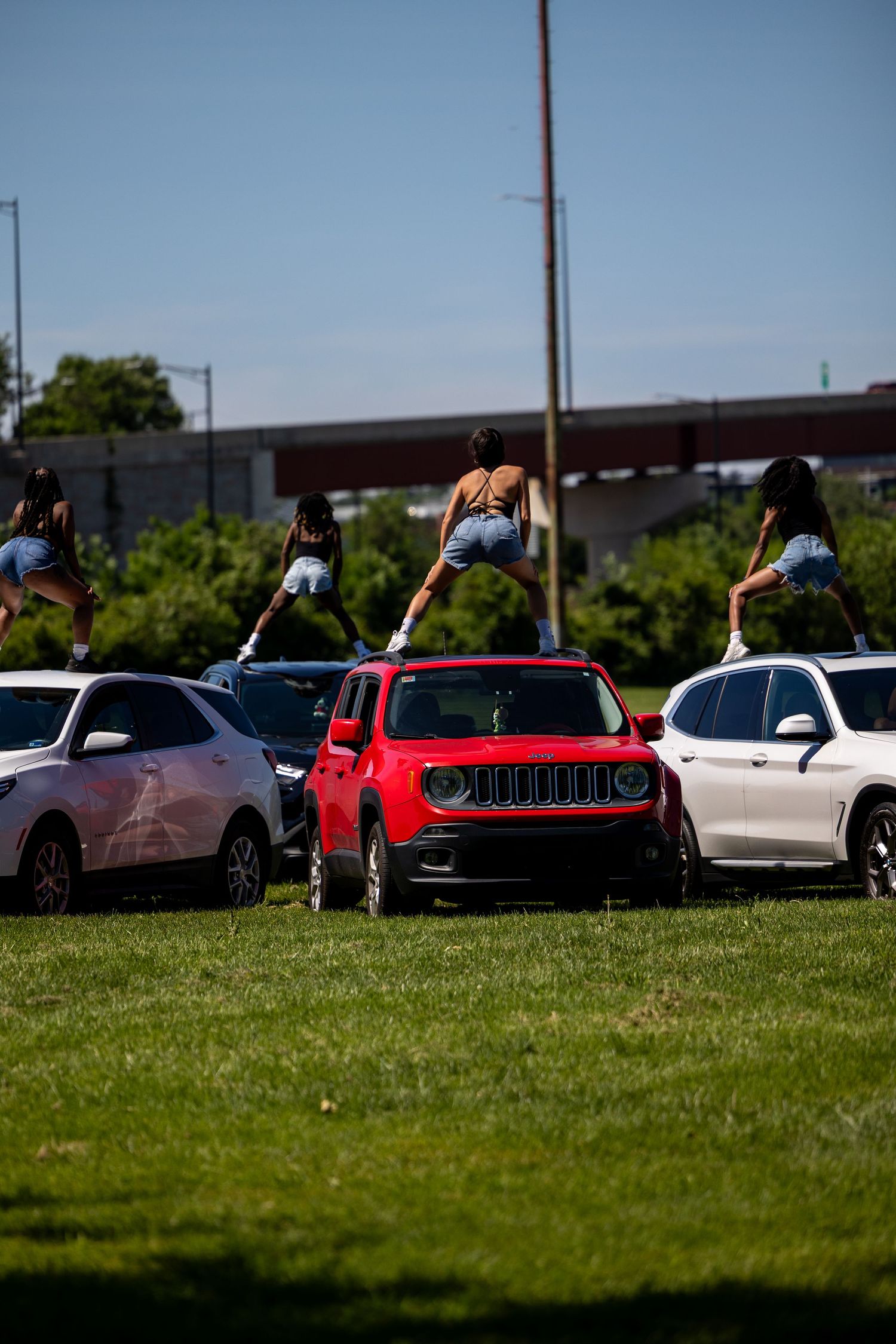Three women in denim shorts standing on cars in a parking lot on a sunny day with a red Jeep Renegade in center.