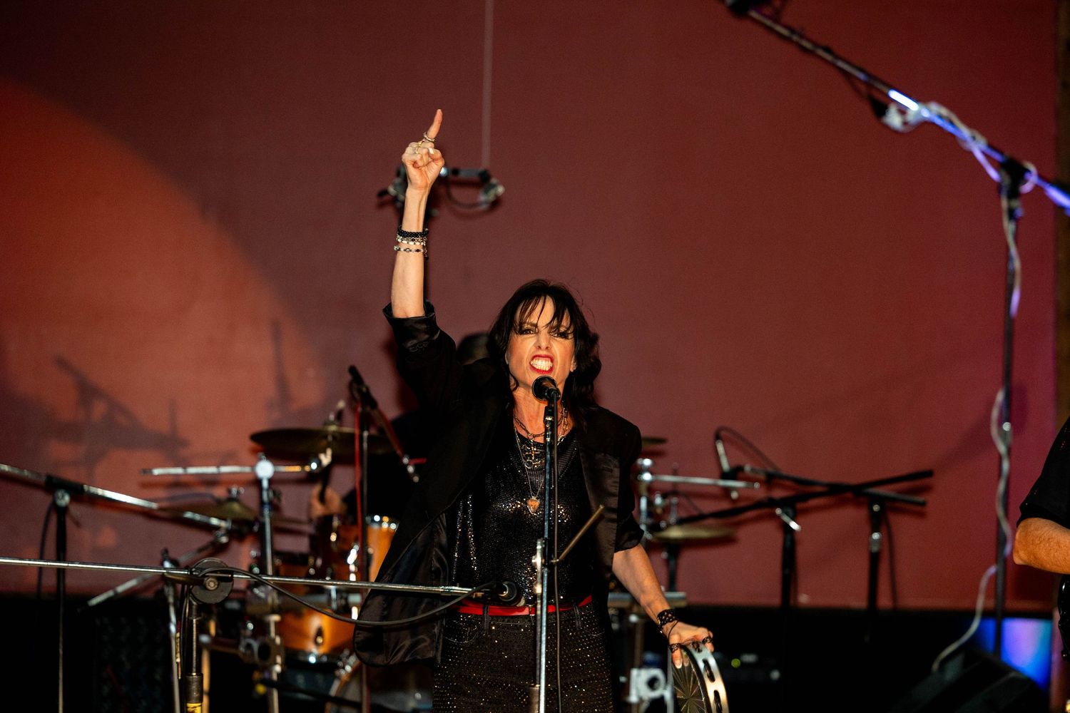 Female rock singer performing on stage, raising one finger while singing into a microphone with drums visible behind her.