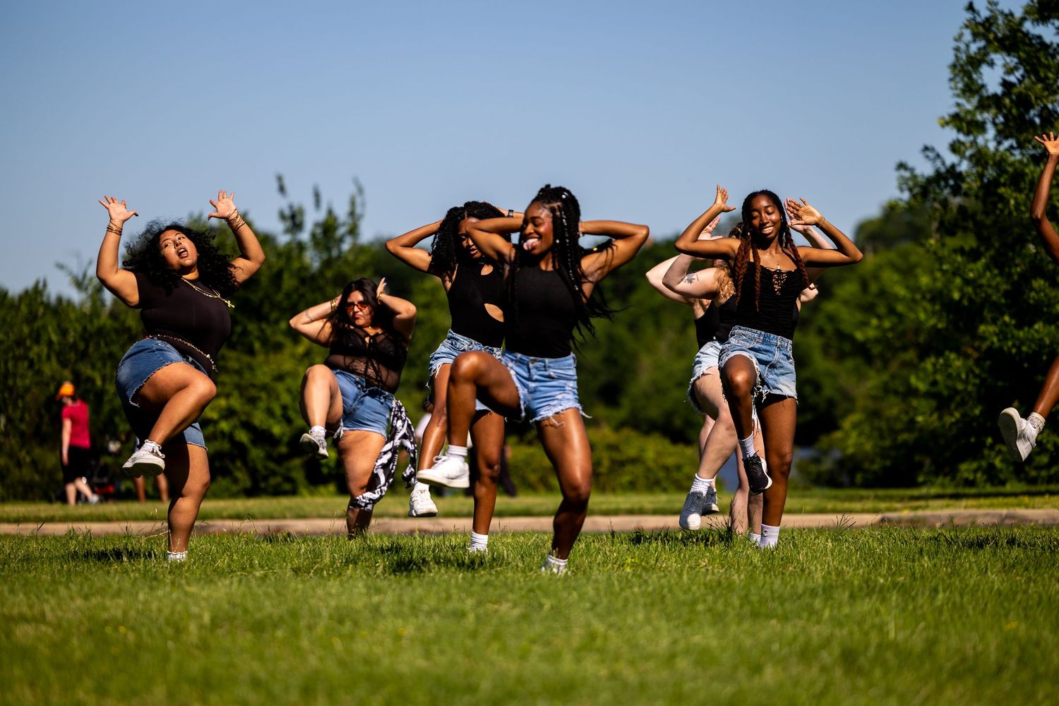 Group of young women dancing energetically outdoors on green grass under a clear blue sky on a sunny day.