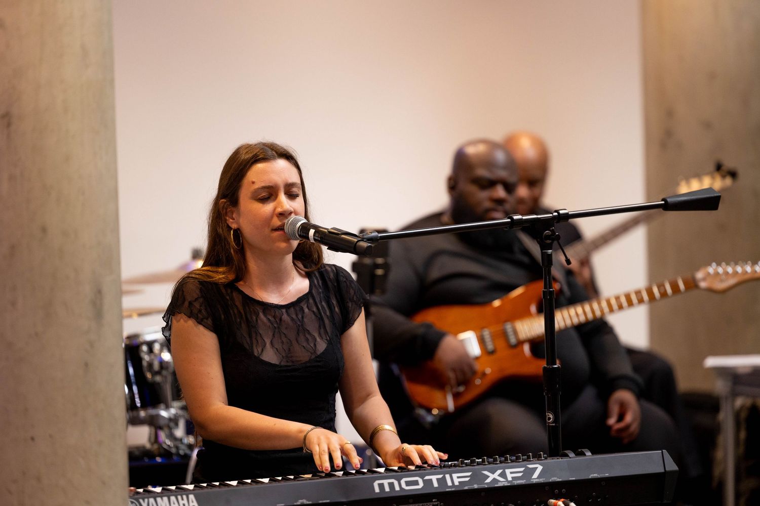 Woman playing Yamaha Motif XF7 keyboard and singing into microphone with guitarist performing in background.