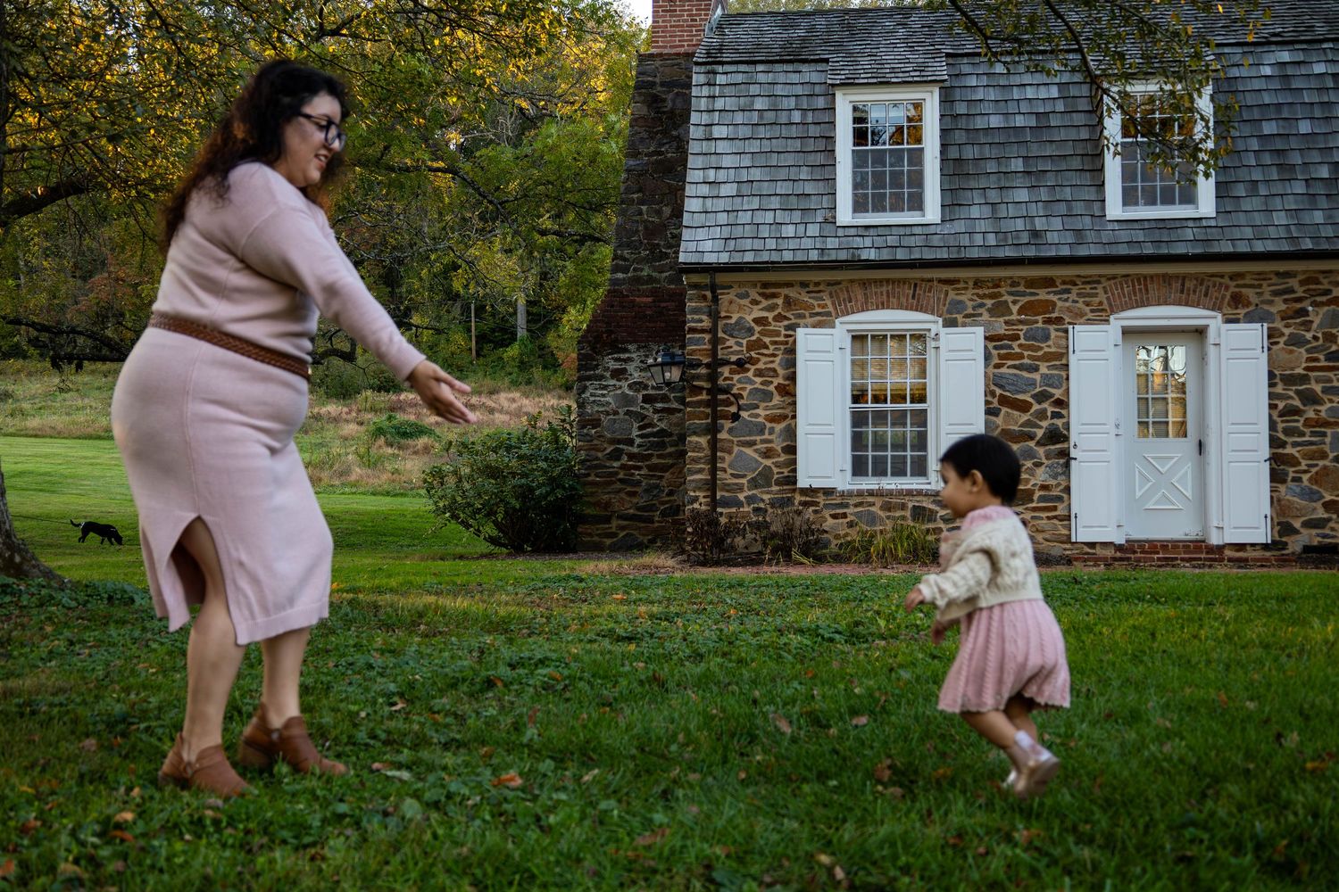 Woman playing with toddler girl in pink dress on green lawn outside historic stone house.