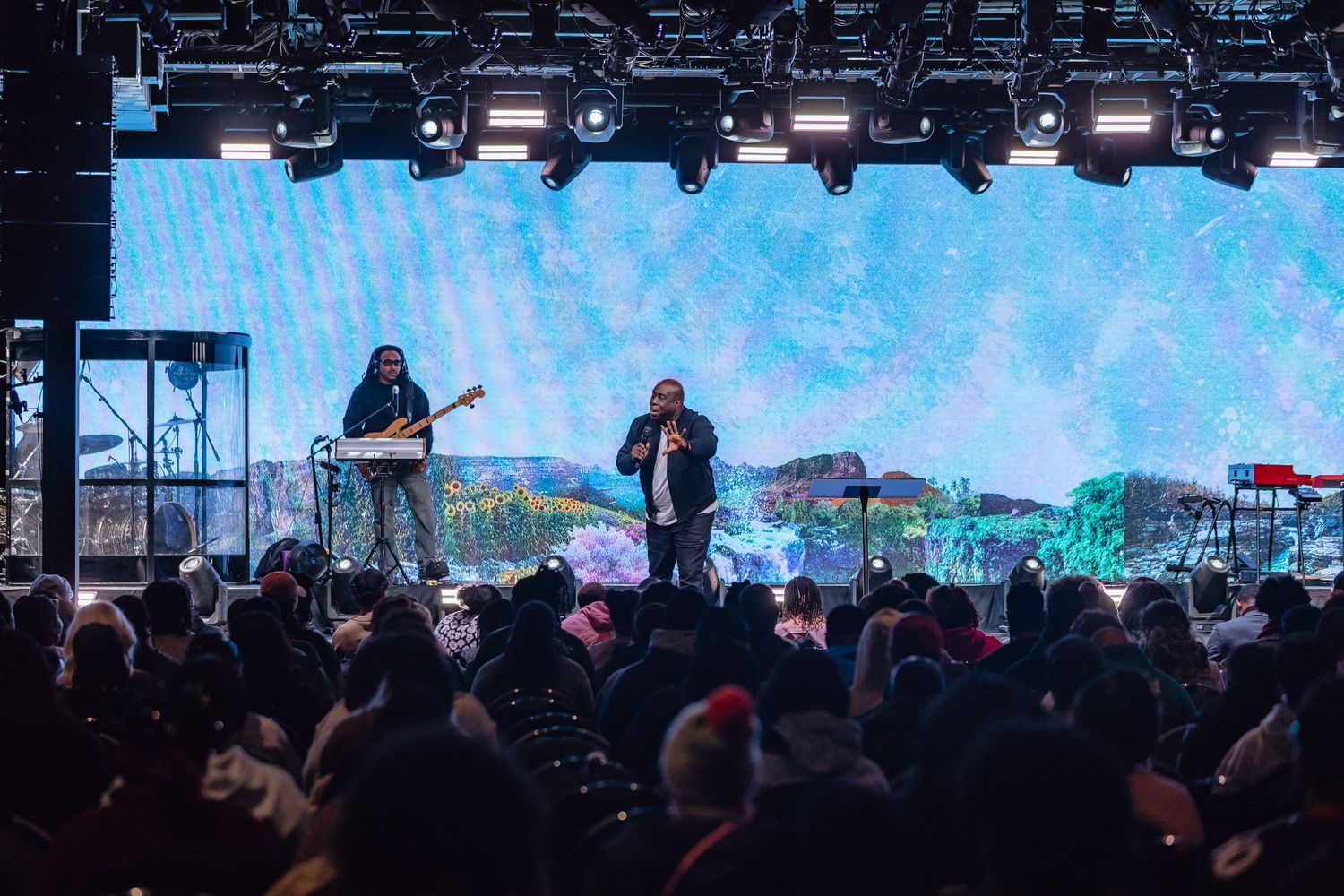 Concert crowd watching performers on stage with bright blue lighting and video screens in the background.
