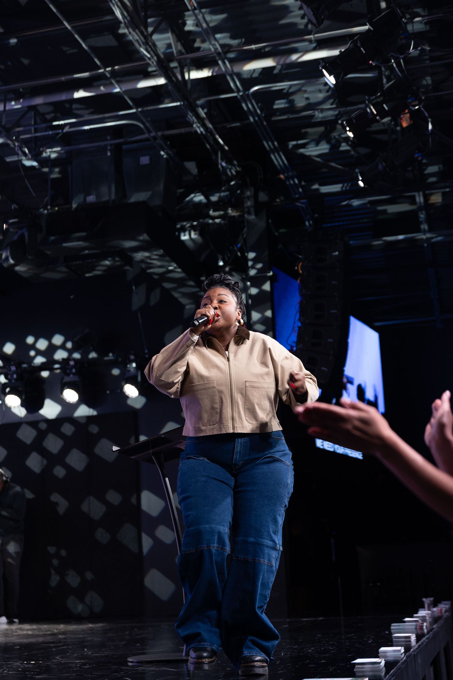 A performer in a beige shirt and blue jeans gestures dramatically on a dark stage under theatrical lighting.