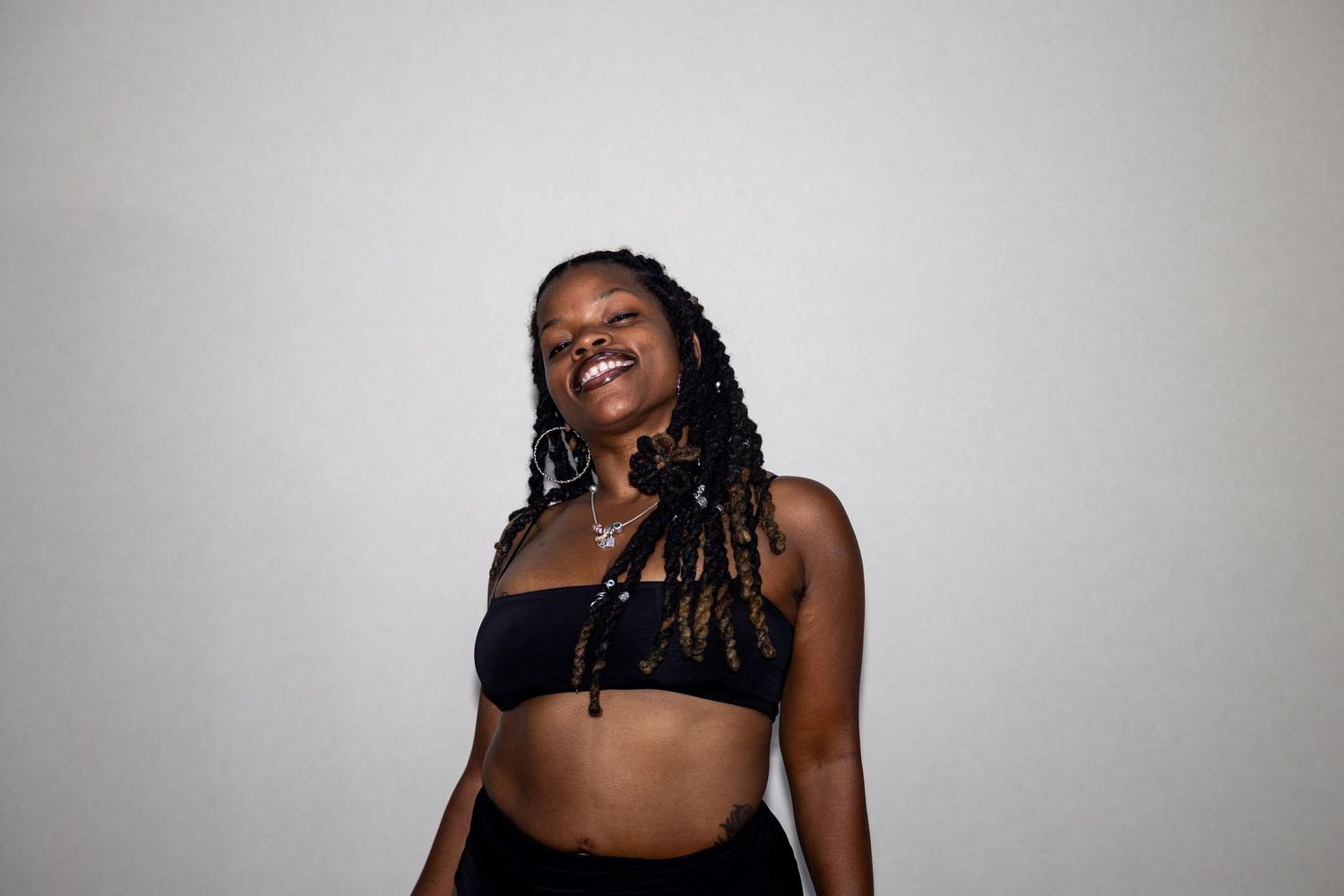 A joyful woman with braided locs wearing a black crop top laughs with head tilted back against a white background.