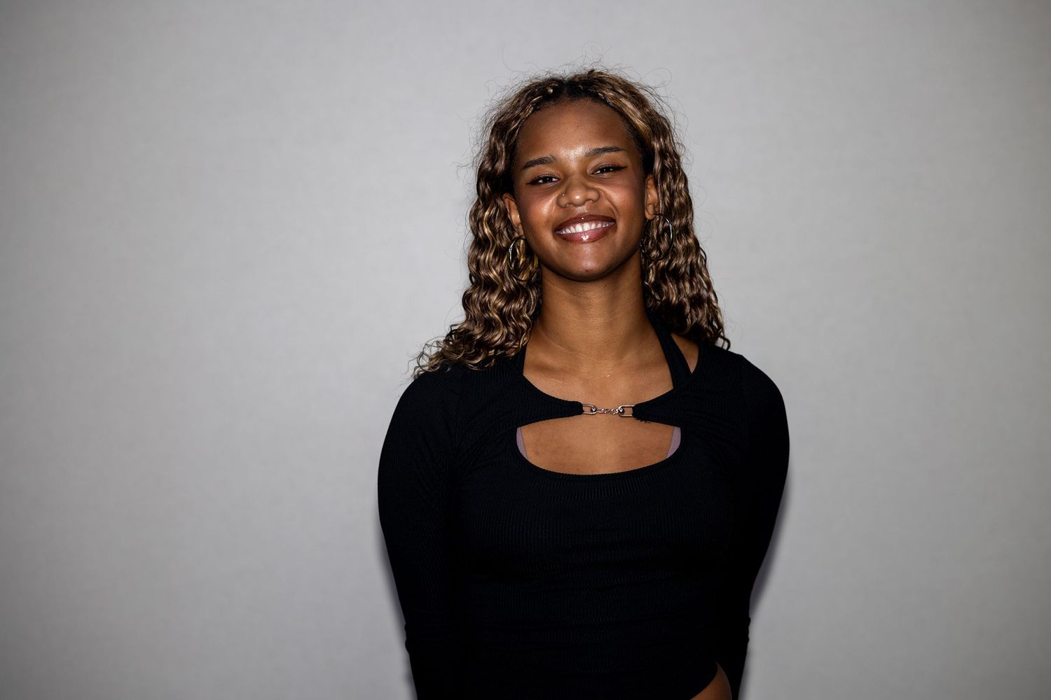 A young woman with voluminous curly hair smiles brightly wearing a black long-sleeve cutout crop top.