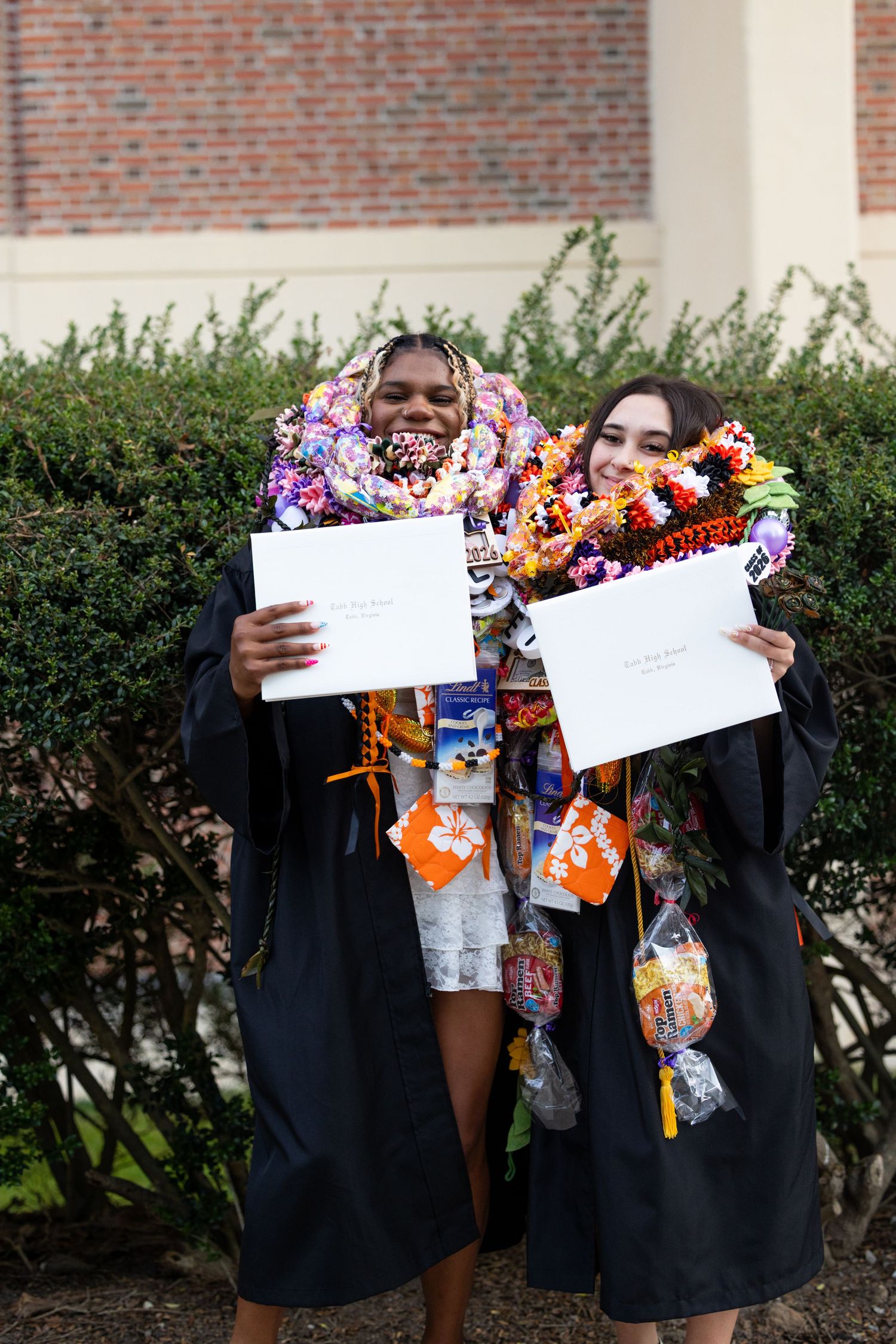 Two graduates in black gowns holding diplomas, adorned with colorful leis and candy necklaces, posing outdoors after commencement.