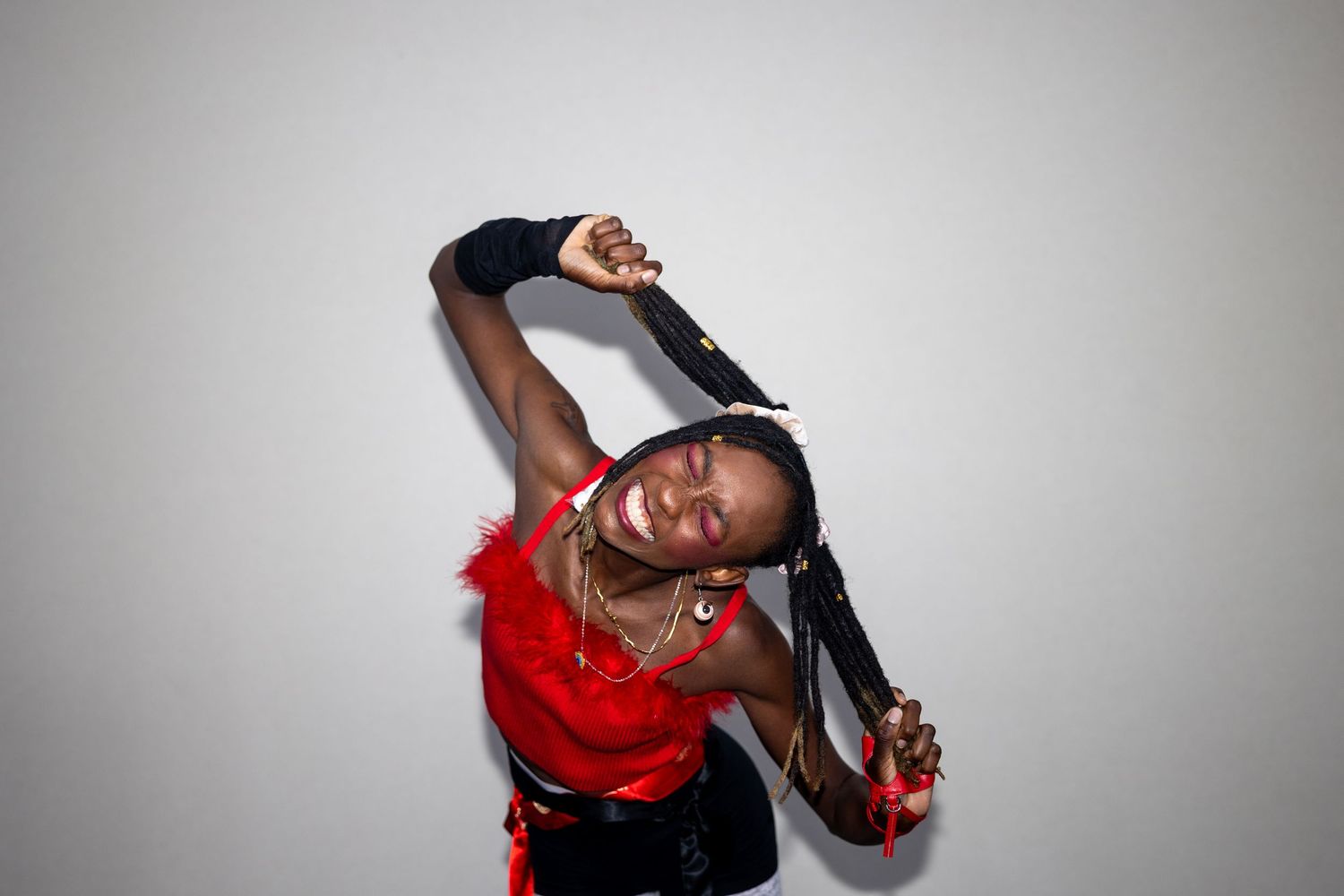 Dynamic performer in red feathered crop top and black gloves posing dramatically while holding her braided hair.