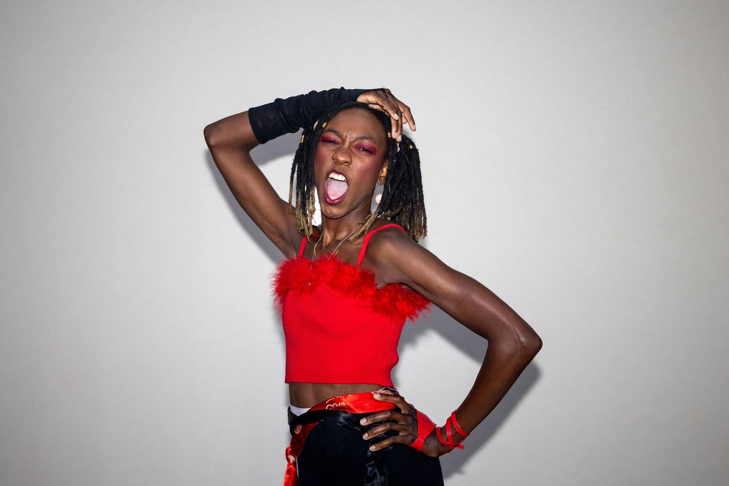 Woman in red crop top and black gloves poses energetically against white background with expressive open mouth.