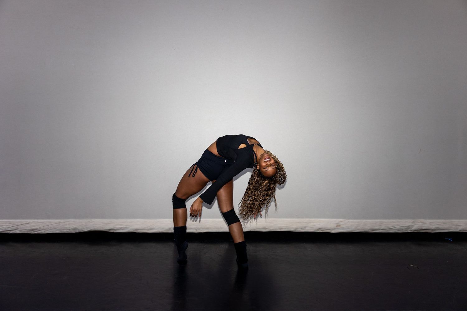Woman in black outfit performing a dramatic backbend dance move on a dark floor against a gray wall.