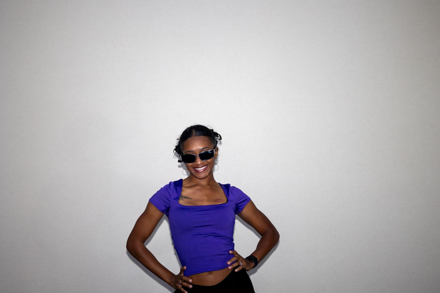 Cheerful woman in purple top and sunglasses smiling with hands on hips against white wall.