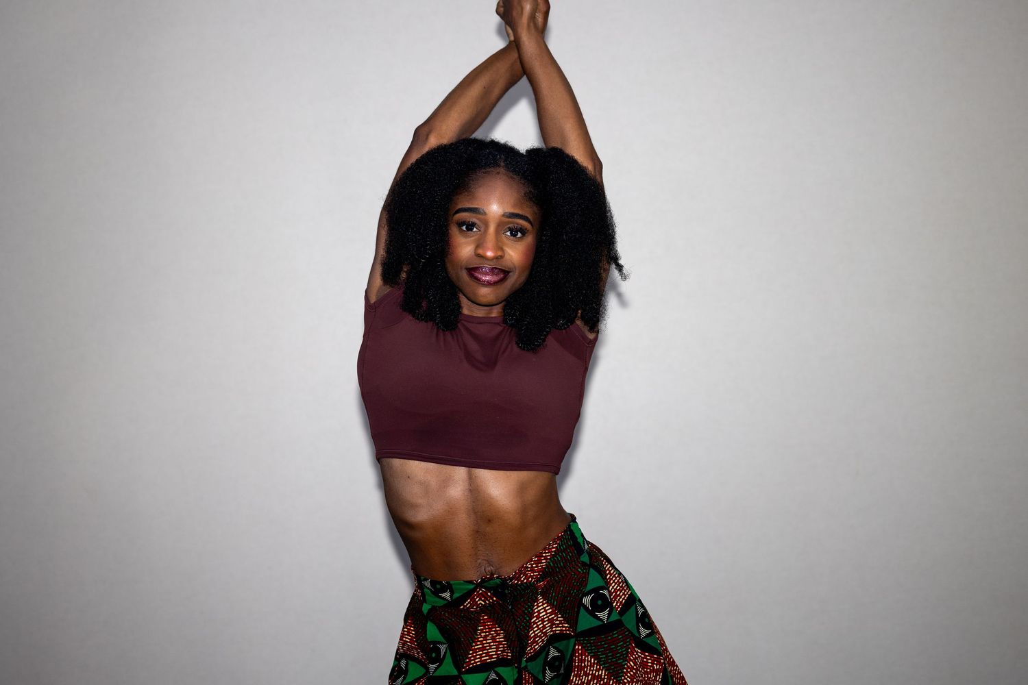 Young Black woman in maroon crop top and African print skirt posing with arms raised overhead against white wall.