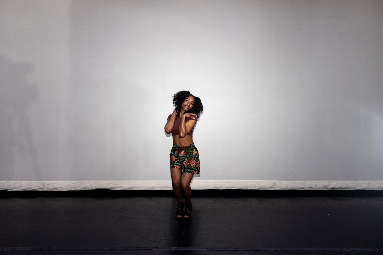 Woman in African print outfit standing with hands near face in expressive dance pose on dark floor.