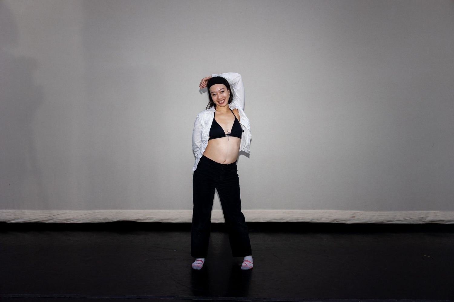 A woman in black pants and white jacket poses in a dance studio.