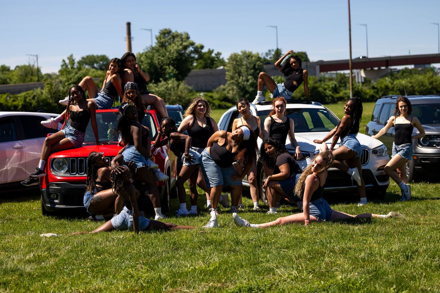 Group of friends posing together outdoors near cars on a sunny day, smiling and celebrating.