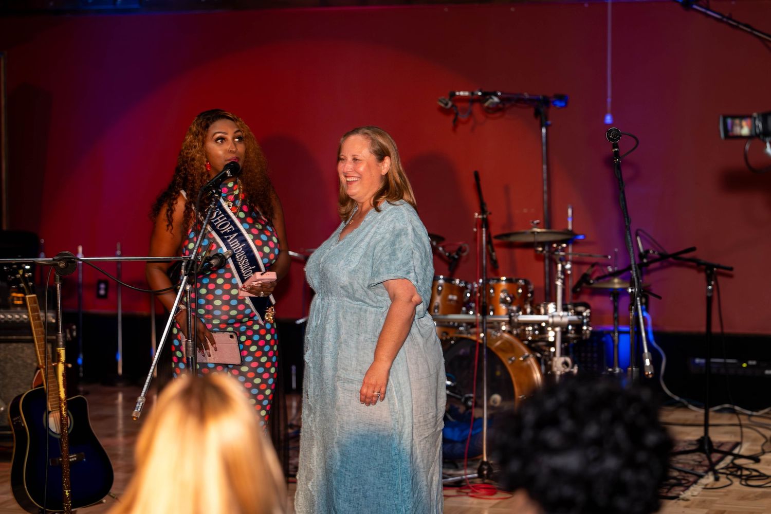 Two women on stage at a live music event, one wearing a sash, with drums and microphones in the background.
