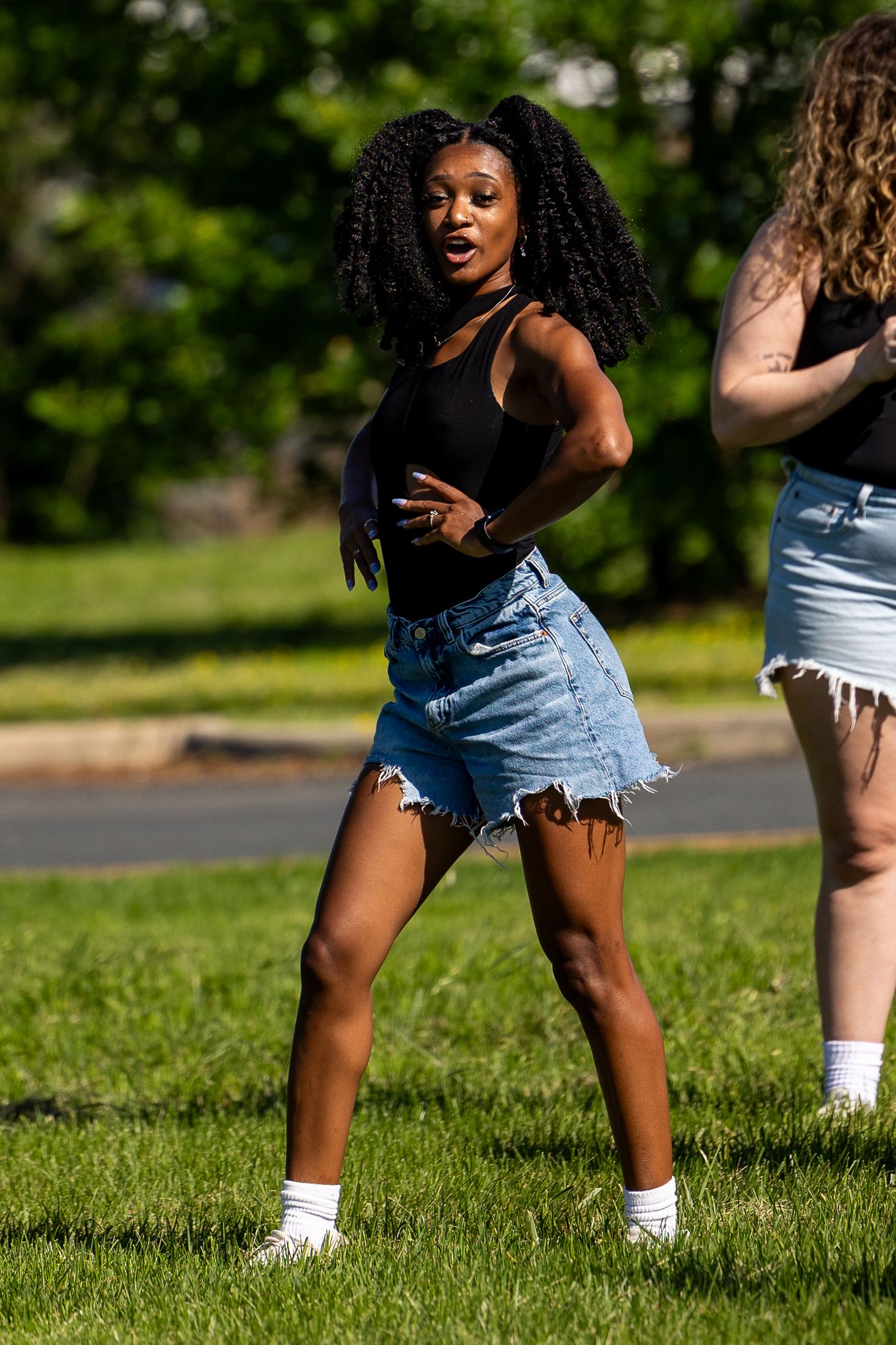 Young Black woman dancing outdoors in denim cutoff shorts and black top on green grass in sunny park.