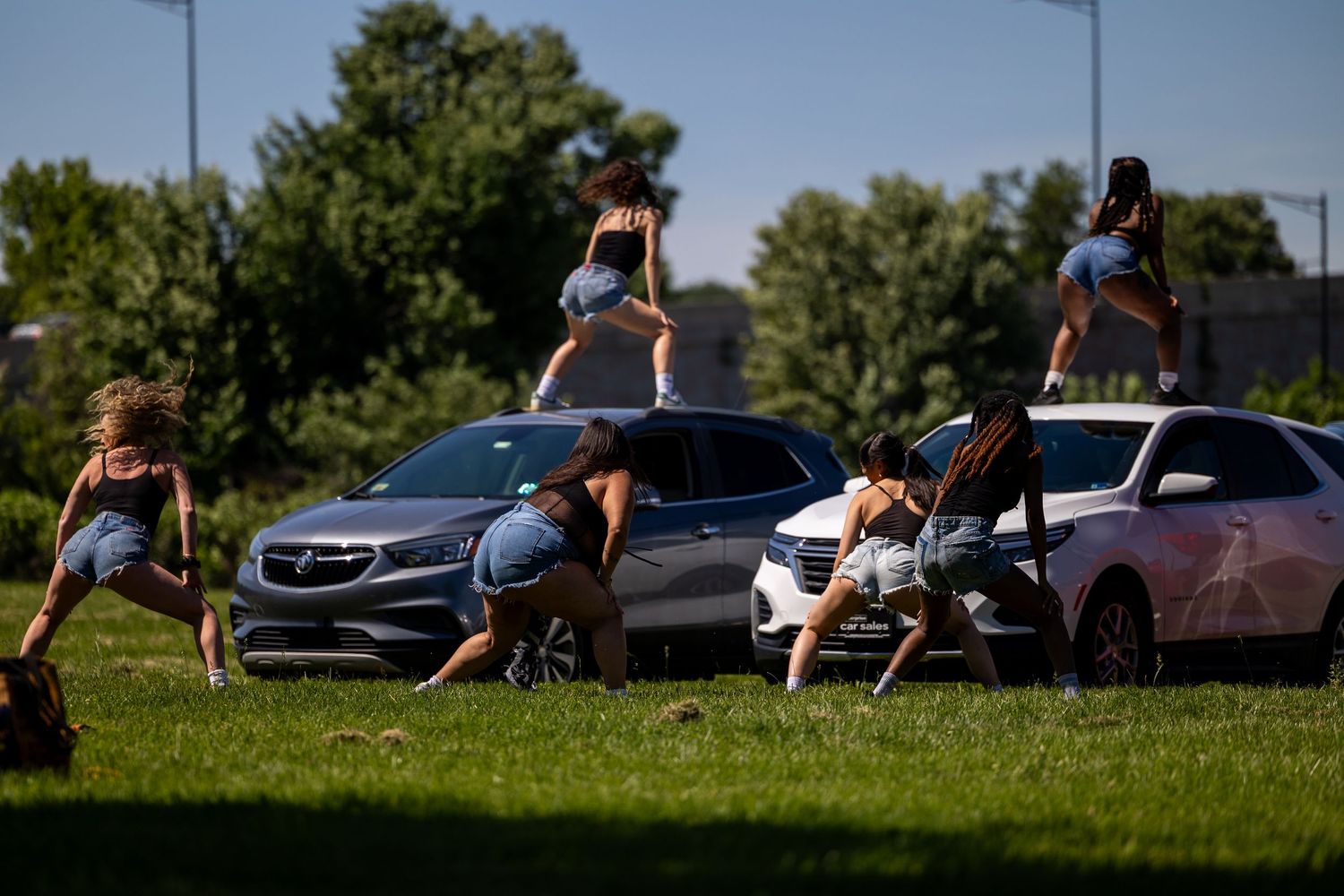 Young women dancing and posing on and around parked SUVs in a grassy outdoor area on a sunny day.