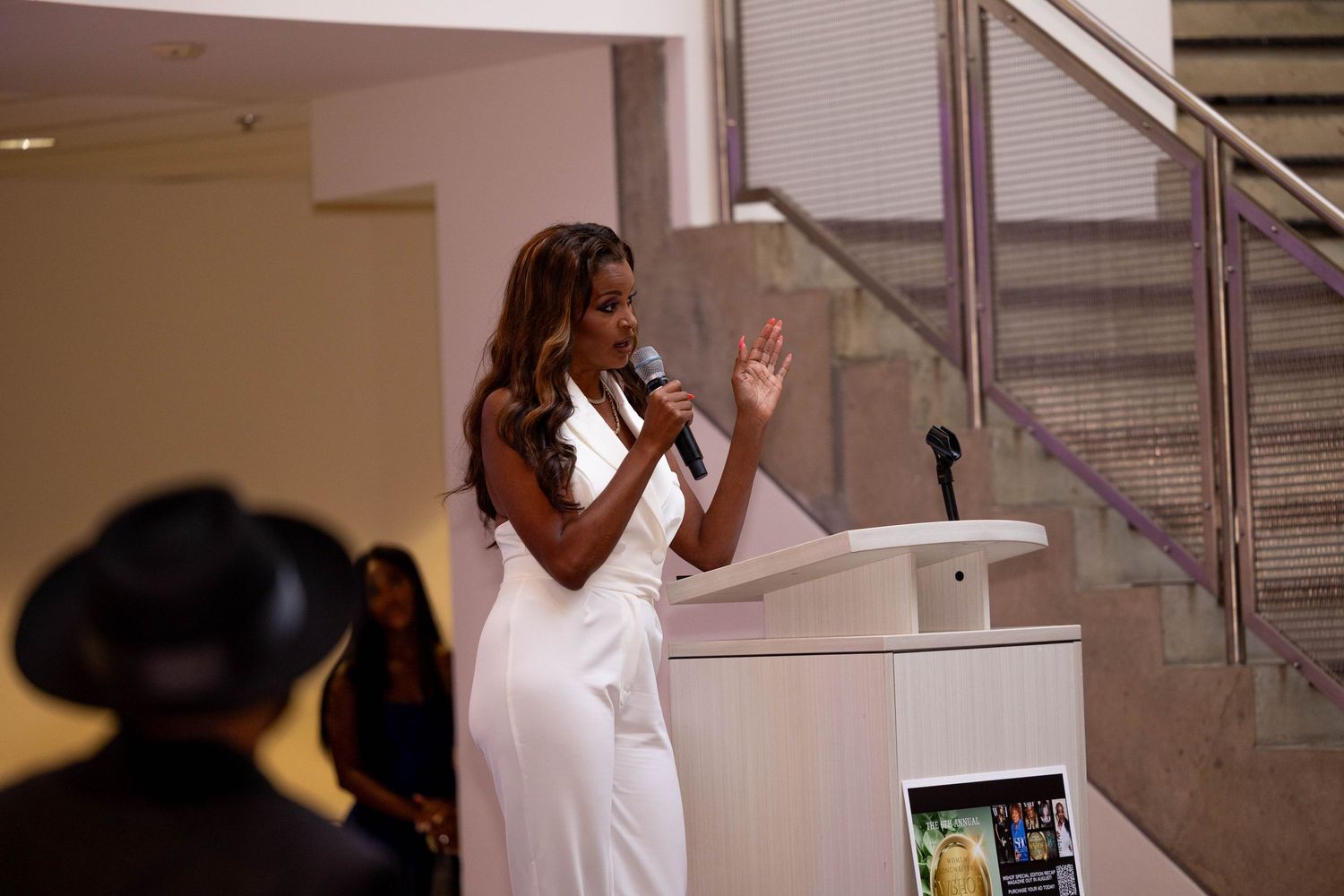 Elegant woman in white dress speaks at podium with microphone at event.