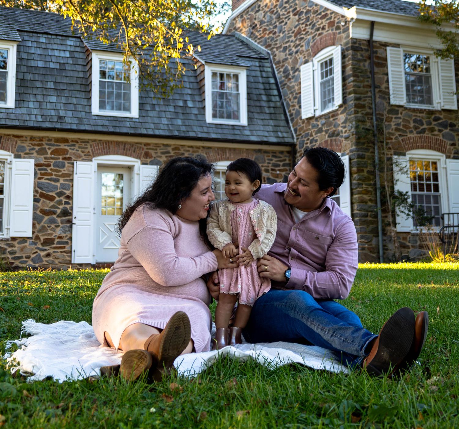 Family of three sitting on blanket in grass outside historic stone home on sunny day.