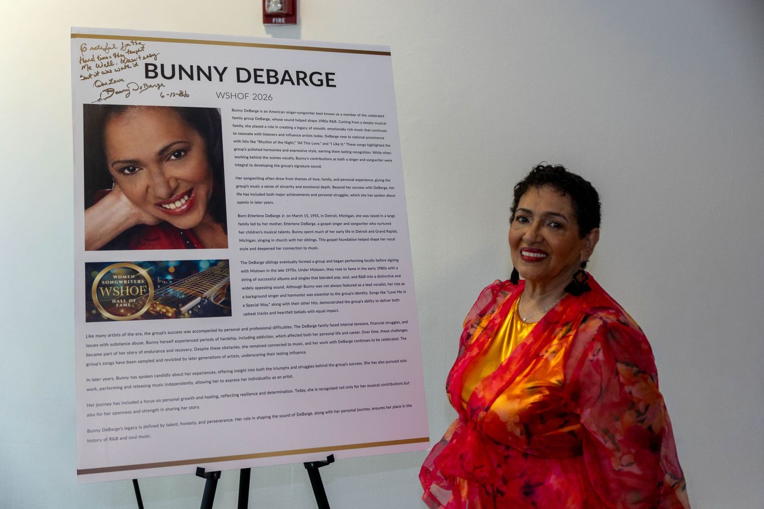 Bunny DeBarge stands next to her name placard display board wearing a vibrant red outfit at an event.