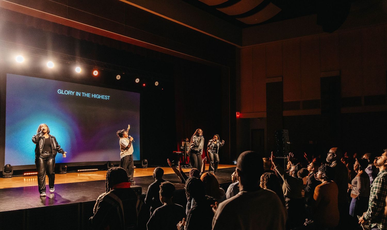 A panoramic view of a dimly lit theater auditorium with stage lighting and audience members visible in silhouette.