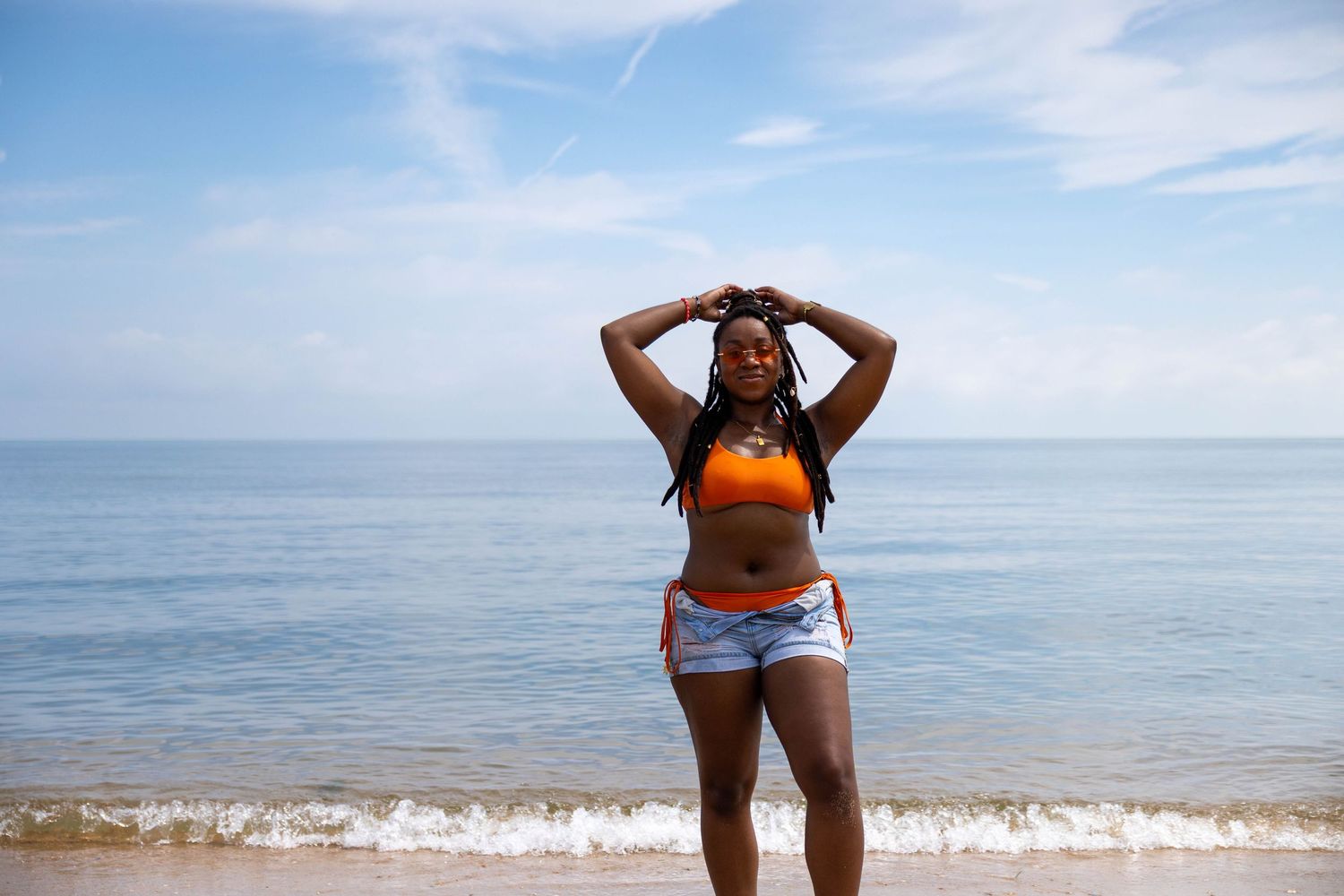 Young woman in orange crop top and denim shorts posing with hands on head at sunny beach shoreline.